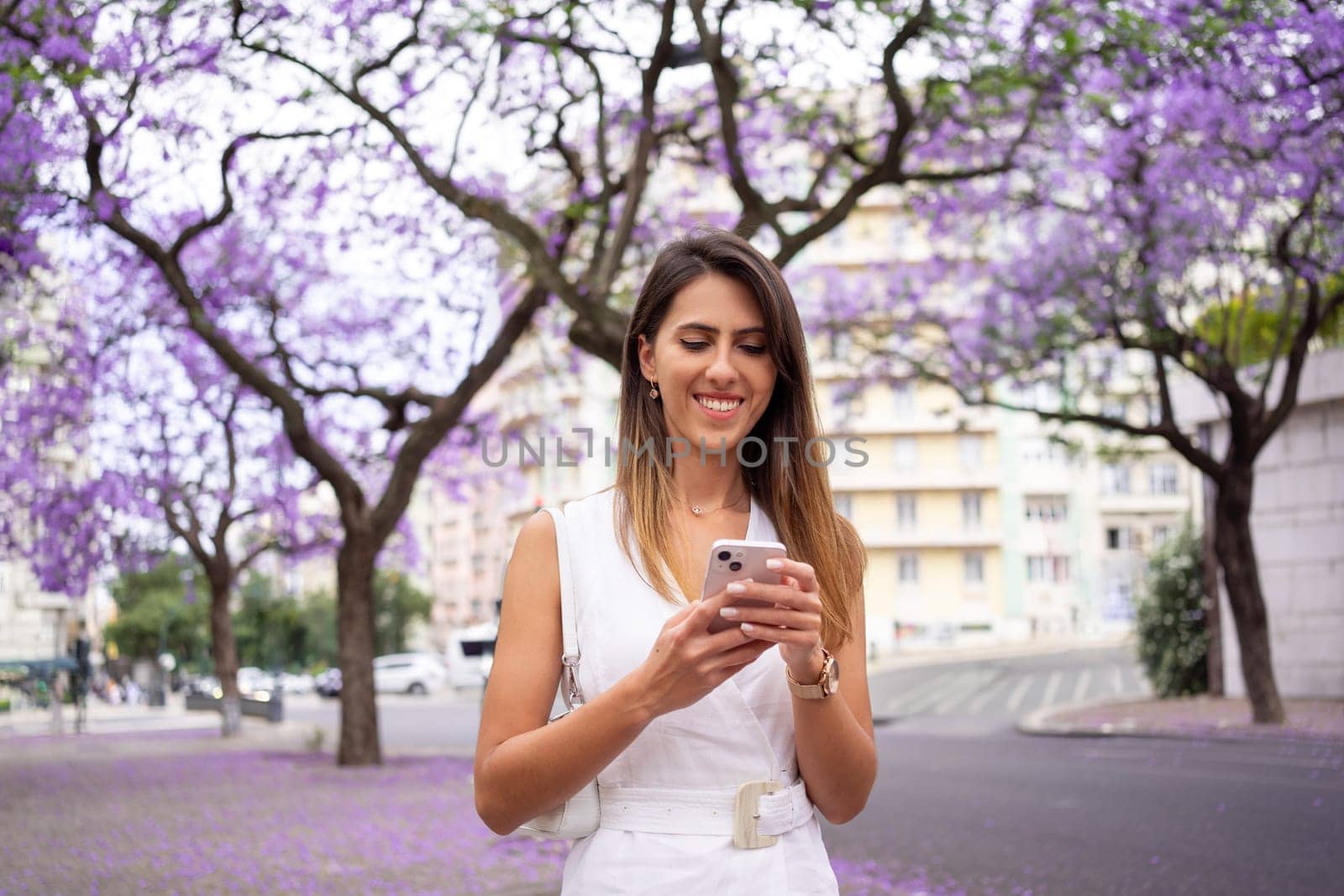 Smiling woman using smartphone near violet Jacaranda trees in city by andreonegin