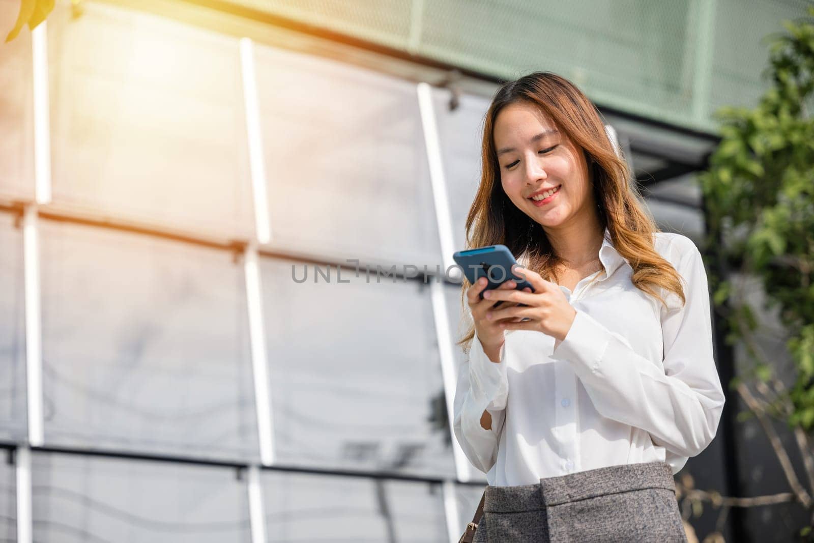 Asian businesswoman holding mobile phone texting message outside of office by Sorapop