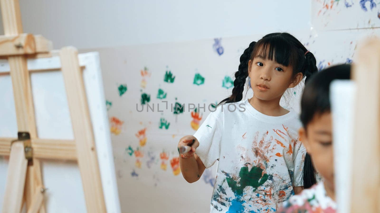 Playful student painted or draw canvas at stained wall in art lesson. Asian girl wearing white shirt with stained color while standing at stained wall with hand print. Creative activity. Erudition.