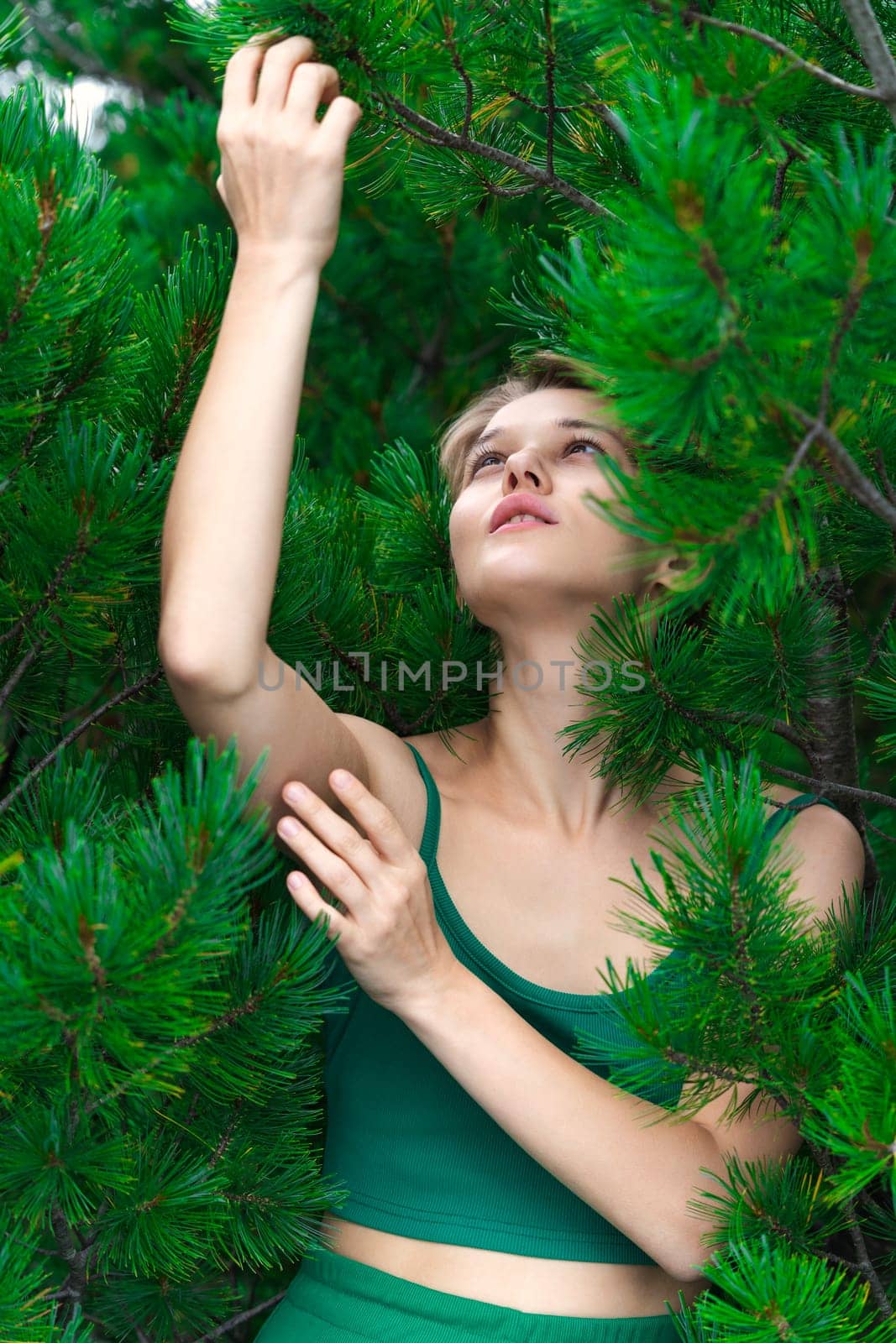 Nosy blonde woman pulls both hands up and looking up, posing against backdrop of an evergreen shrub of Pinus Pumila. Cute Caucasian adult female dressed in green crop top. Part of series