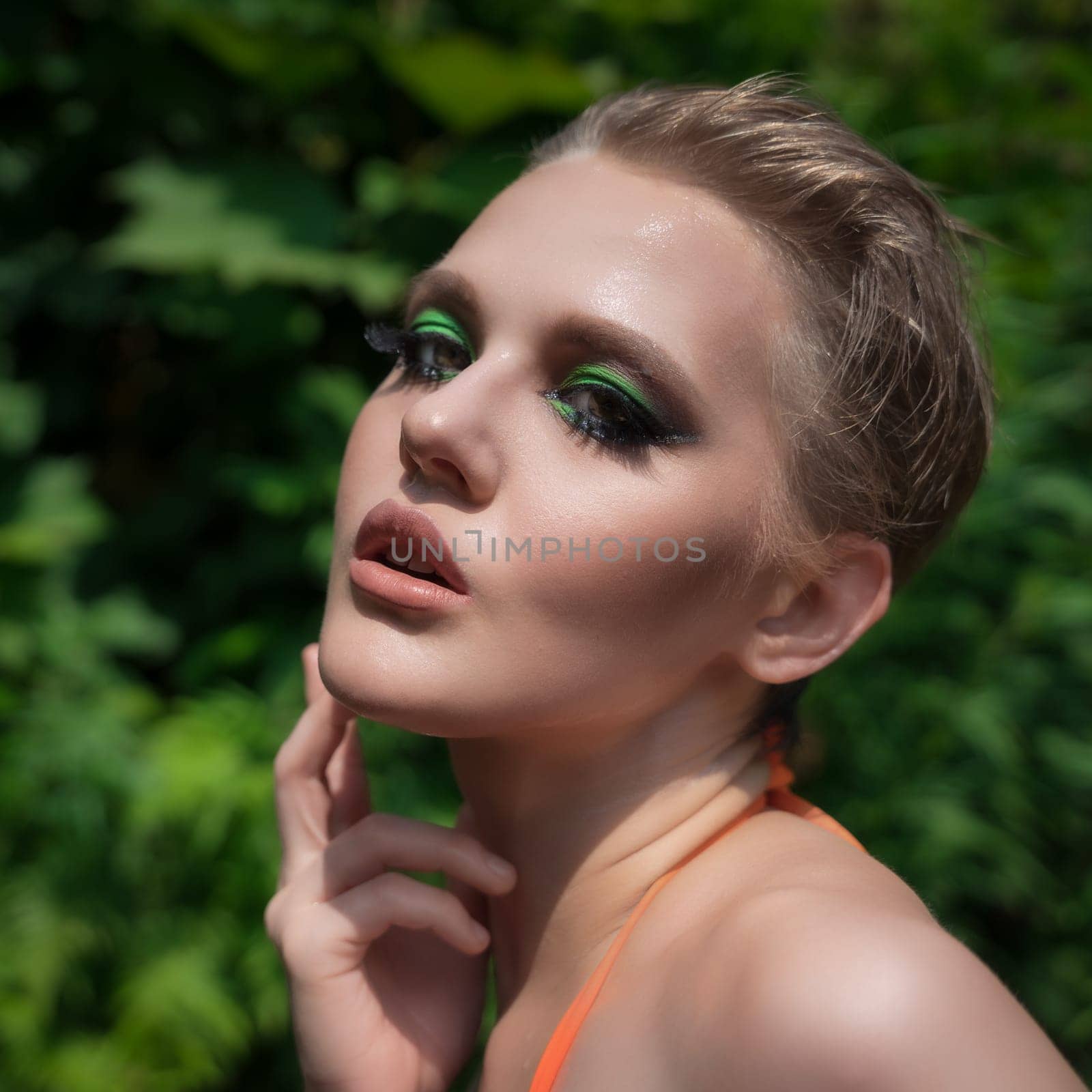 Close-up portrait of blonde woman with short hair, bright make-up against background of green leaves in summer forest. Beauty adult female with sensually parted mouth stares