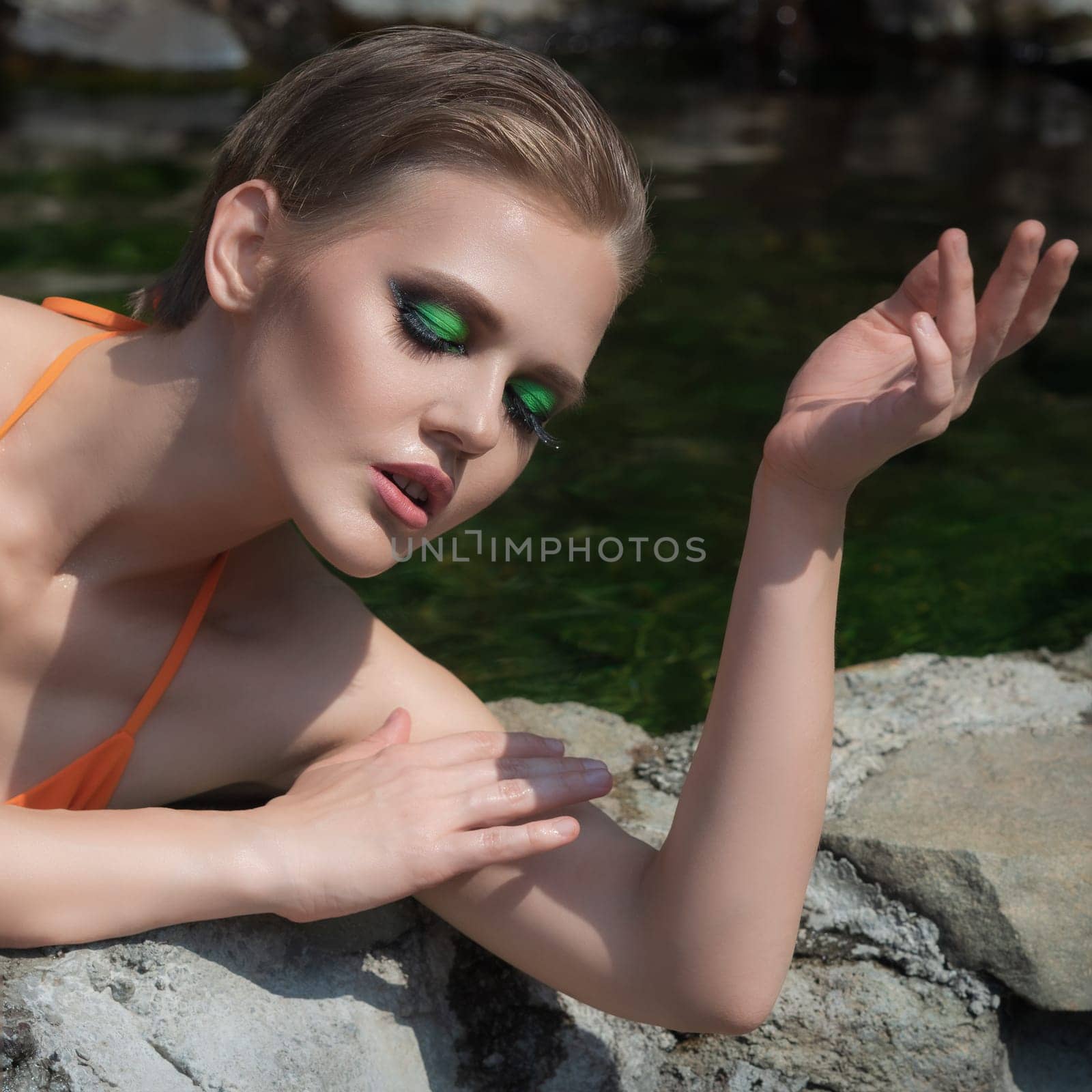 Adult woman with closed eyes and bright makeup poses with her hands on the rocky edge of an outdoor pool filled with natural geothermal water. Fashionable female is dressed in an orange bikini top