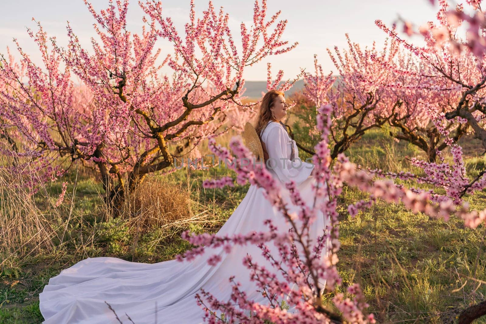 Woman blooming peach orchard. Against the backdrop of a picturesque peach orchard, a woman in a long white dress and hat enjoys a peaceful walk in the park, surrounded by the beauty of nature. by Matiunina