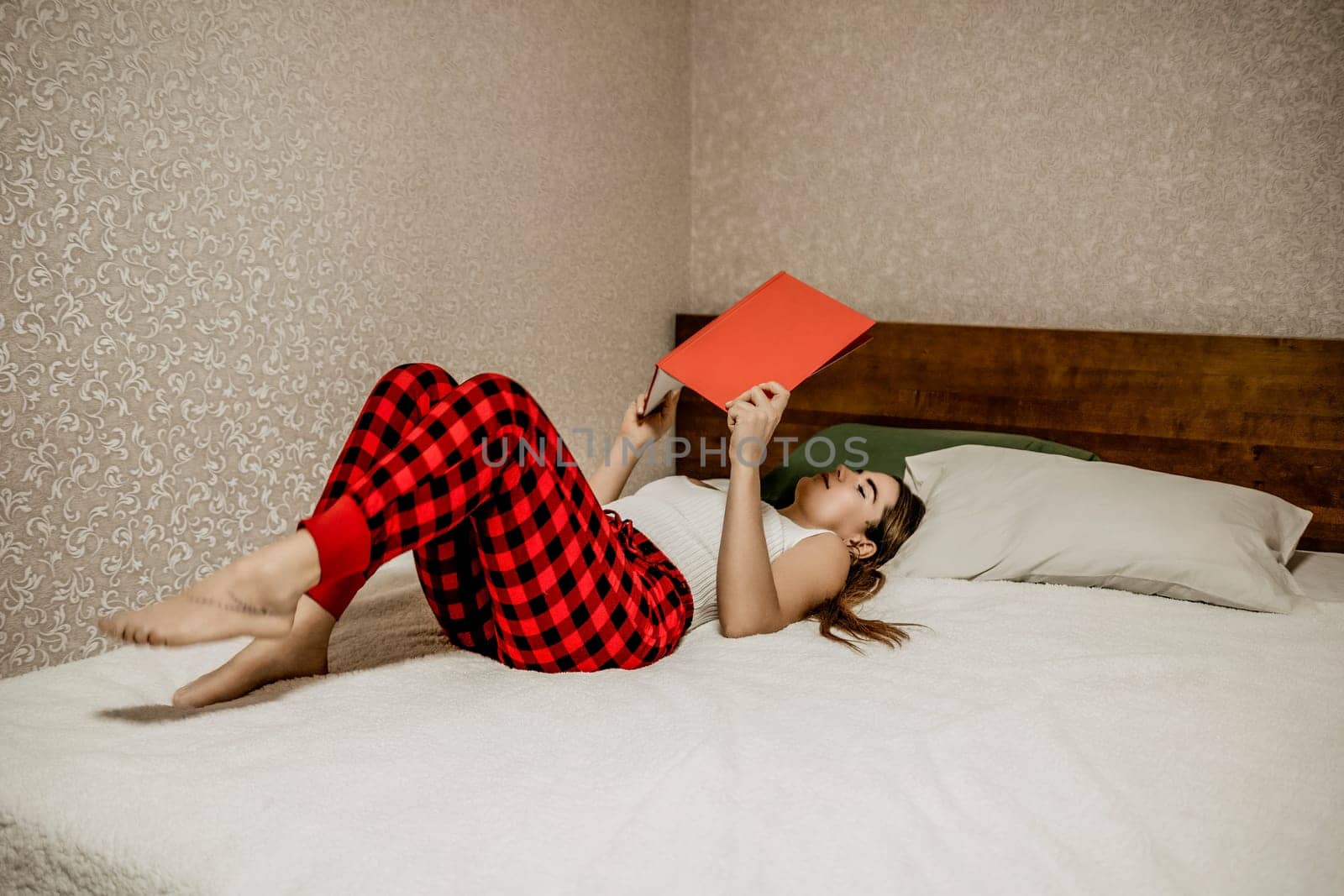 Young brunette woman works and studies from home on a laptop, while lying on her bed in red checkered pants. Balancing work and studies at home