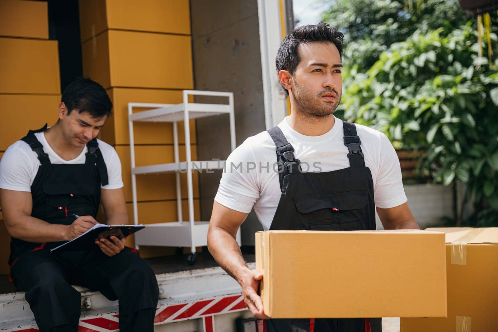 Uniformed workers unload boxes inspecting checklist with a clipboard at the truck. Professional delivery team guarantees efficient relocation and reliable service. Moving day concept by Sorapop
