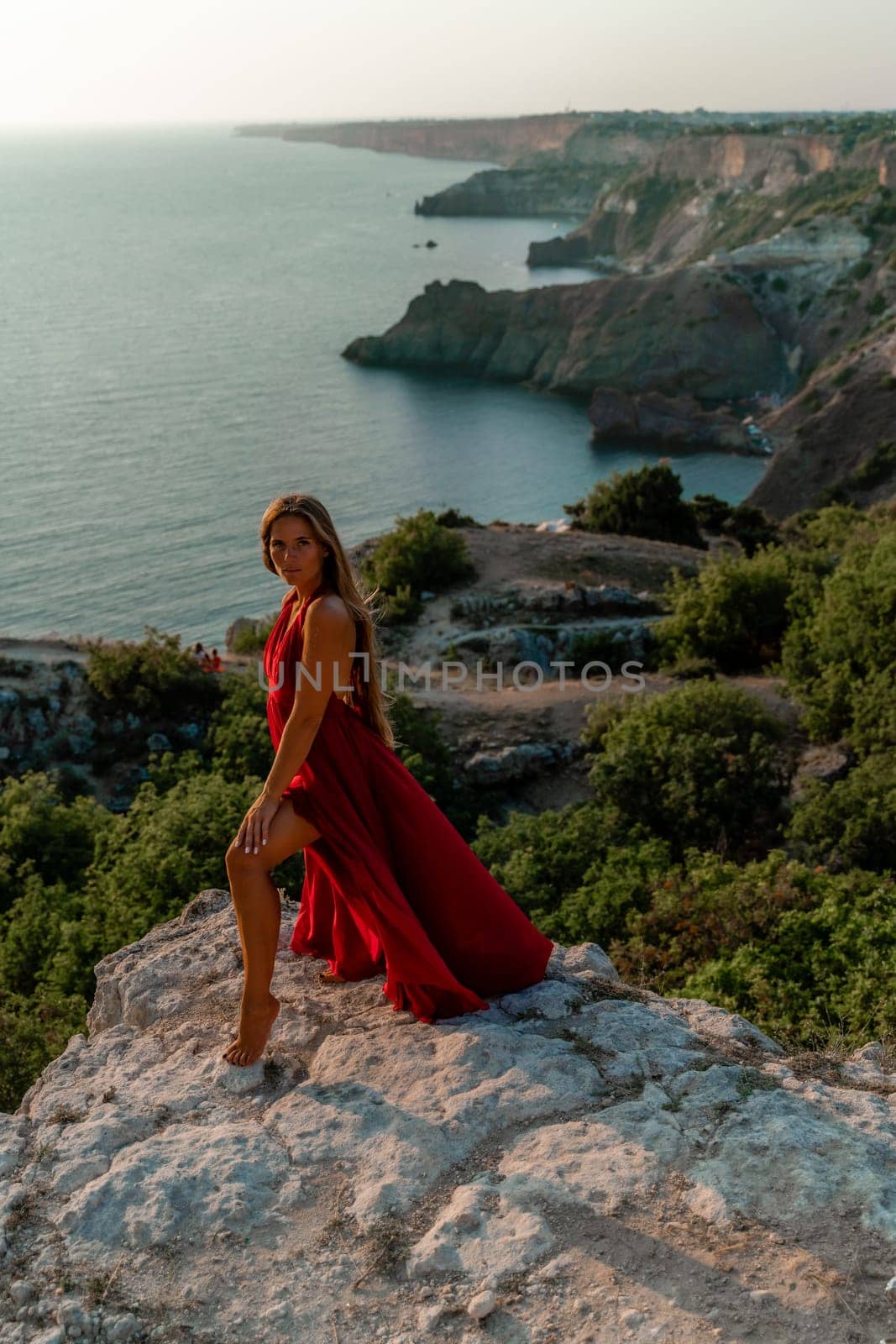 Woman sunset sea red dress, side view a happy beautiful sensual woman in a red long dress posing on a rock high above the sea on sunset. by Matiunina