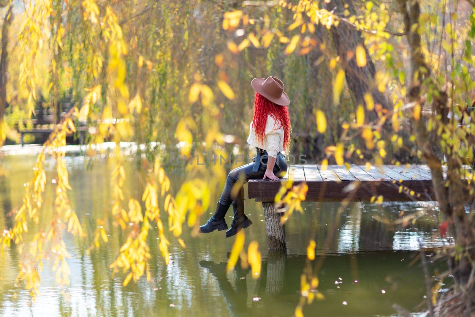 Autumn lake woman. In autumn, she sits by the pond on a wooden pier and admires nature with red hair and a hat. Tourism concept, weekend outside the city