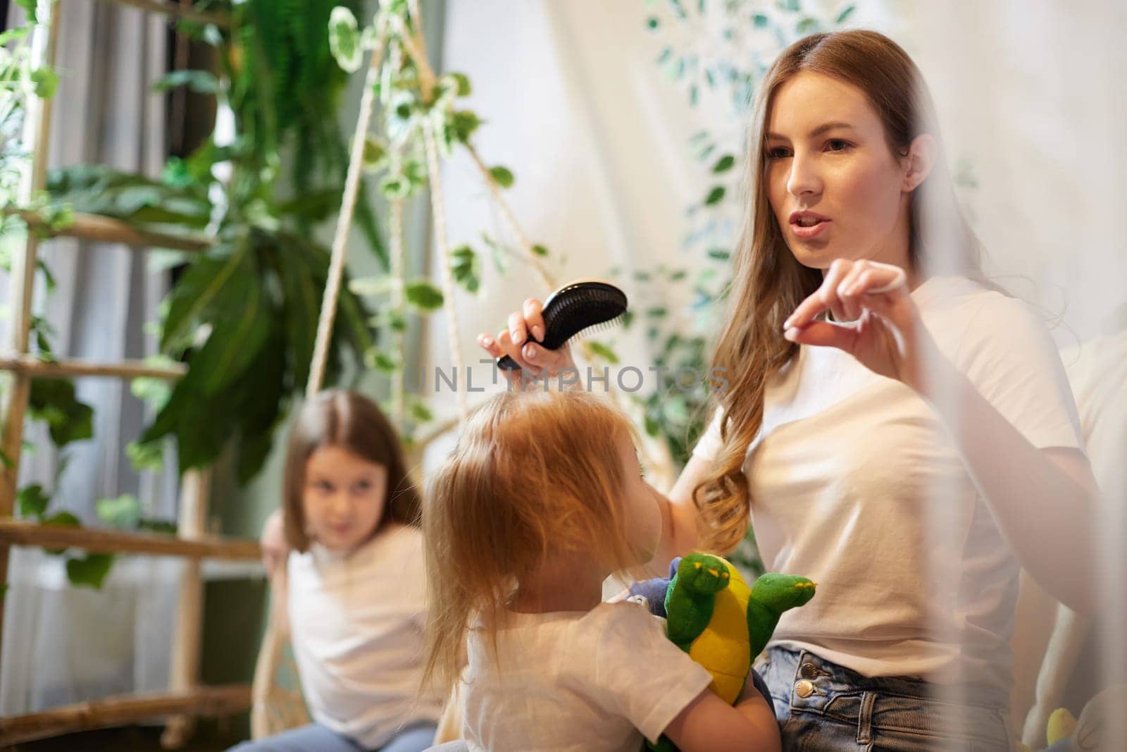 Young loving mom making ponytail to little preschool daughter, adult sister on background in living room. Mother helping child girl with hairstyle