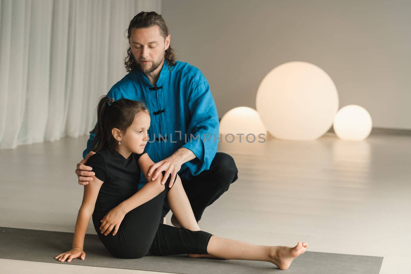 A yoga instructor in training helps a child to do exercises in the gym by Lobachad