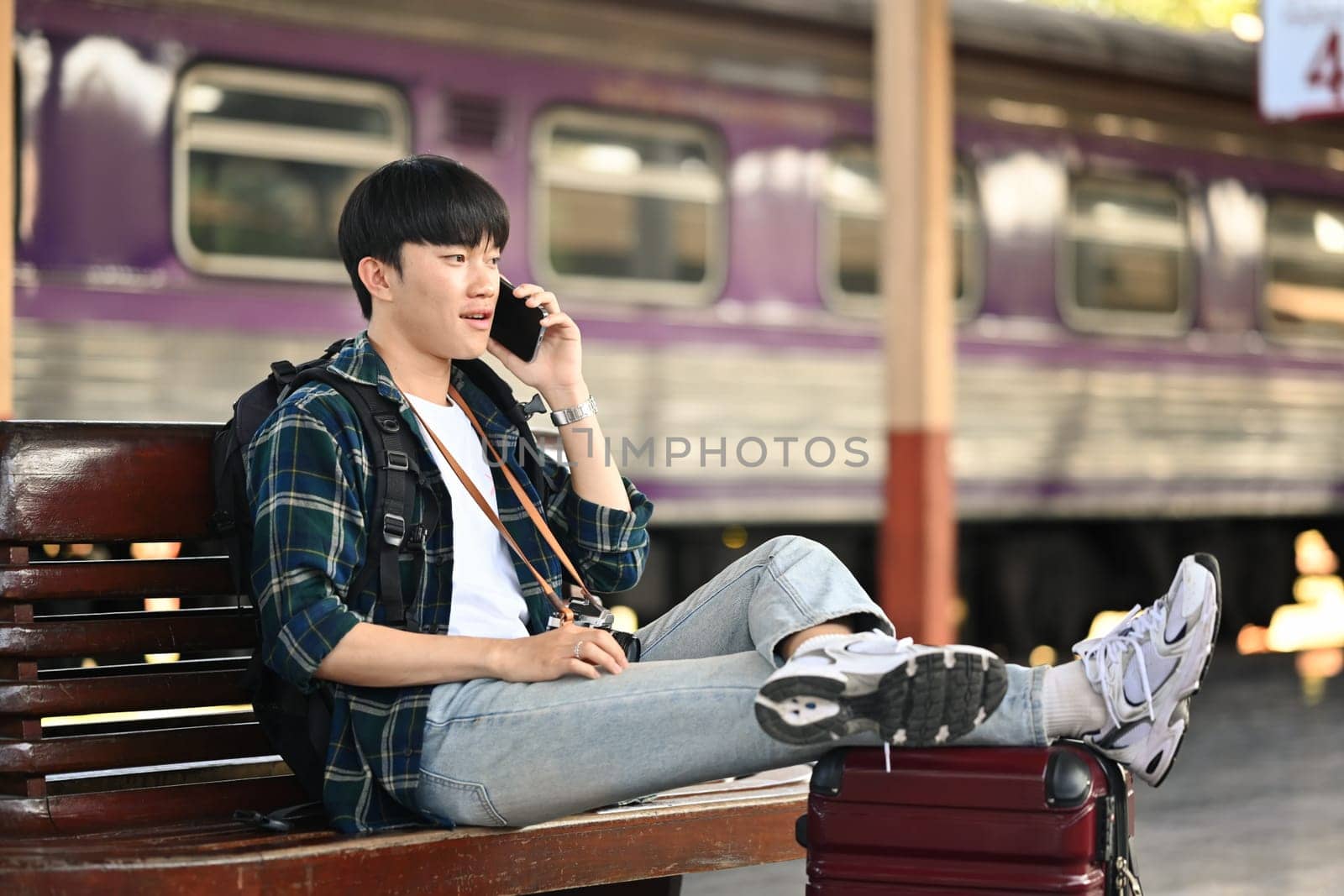 Smiling male tourist talking on mobile phone while waiting train at railway platform. Travel and vacations concept