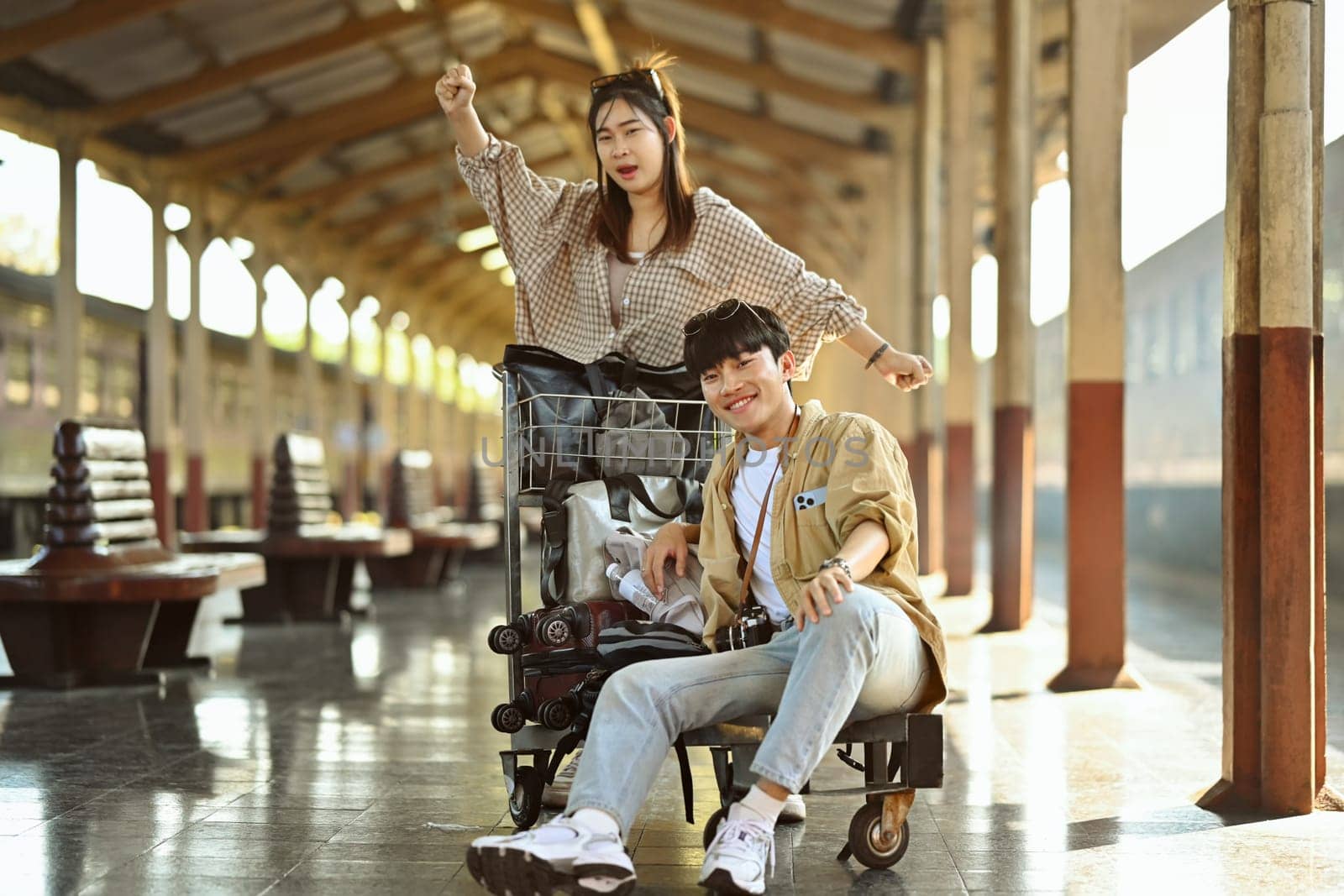 Happy young Asian couple waiting for train in railway station platform. Travel and lifestyle concept