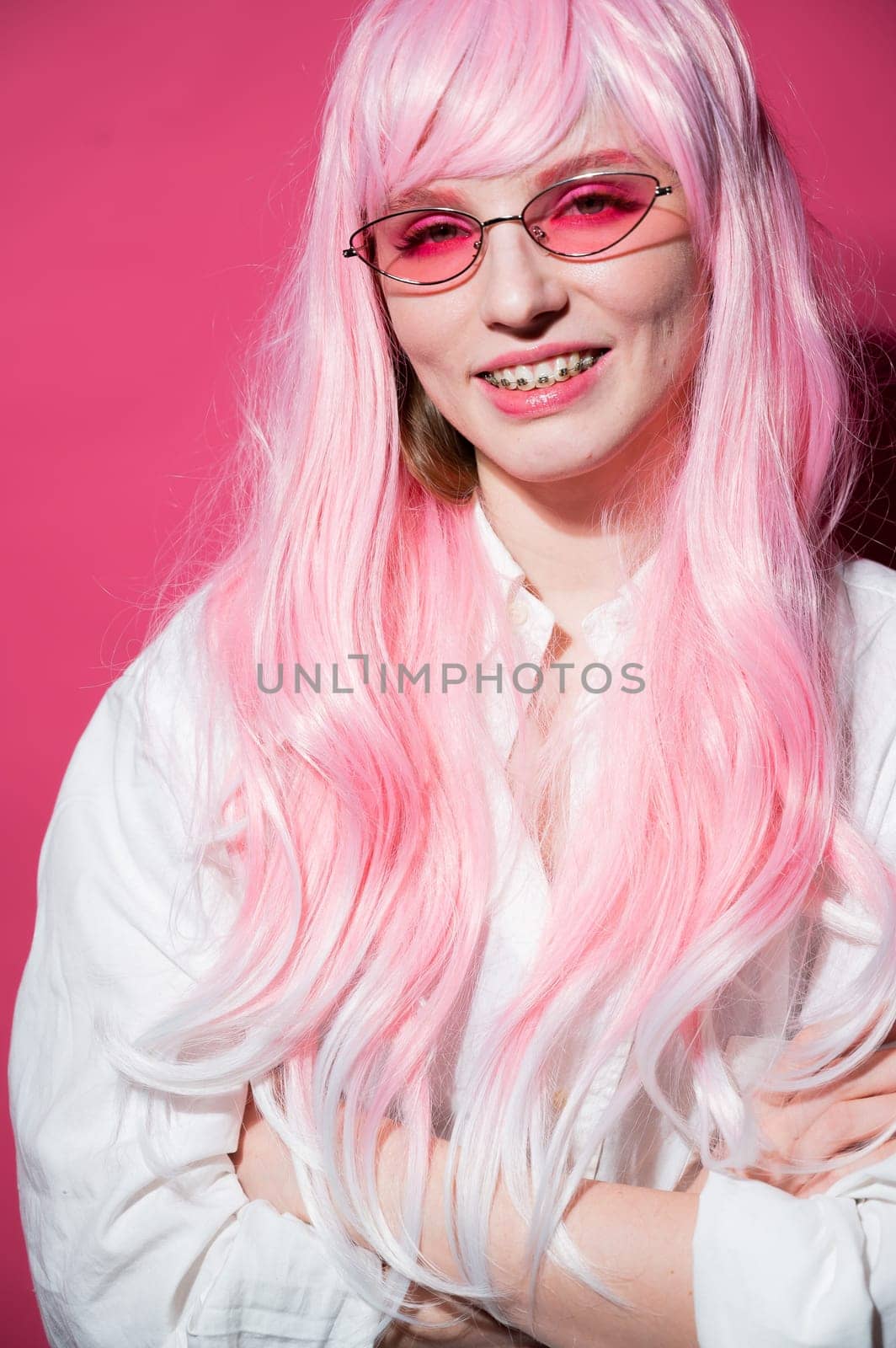 Close-up portrait of a young woman with braces in a pink wig and sunglasses on a pink background. Vertical photo. by mrwed54