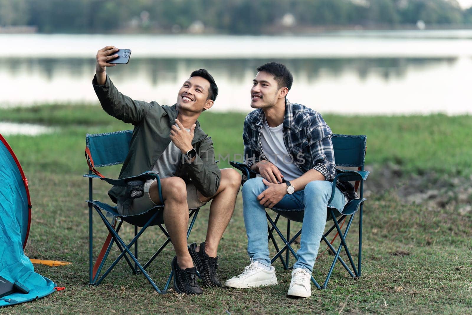 Gay LGBTQIA couple sitting on picnic chair taking selfie together while camping on vacation holiday.