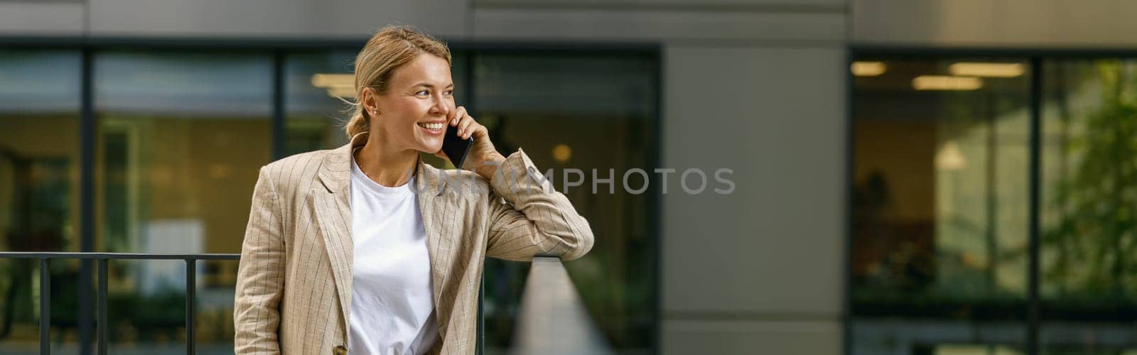 Smiling female manager talking phone standing on modern office building background