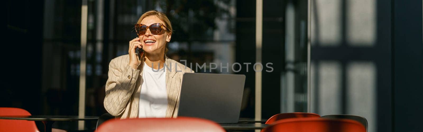Woman manager in sunglasses talking on phone with client while working on laptop in modern coworking by Yaroslav_astakhov