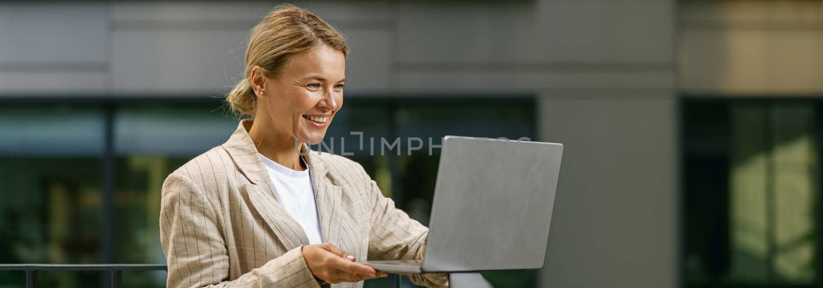 Charming smiling business woman working laptop while standing on modern office terrace by Yaroslav_astakhov