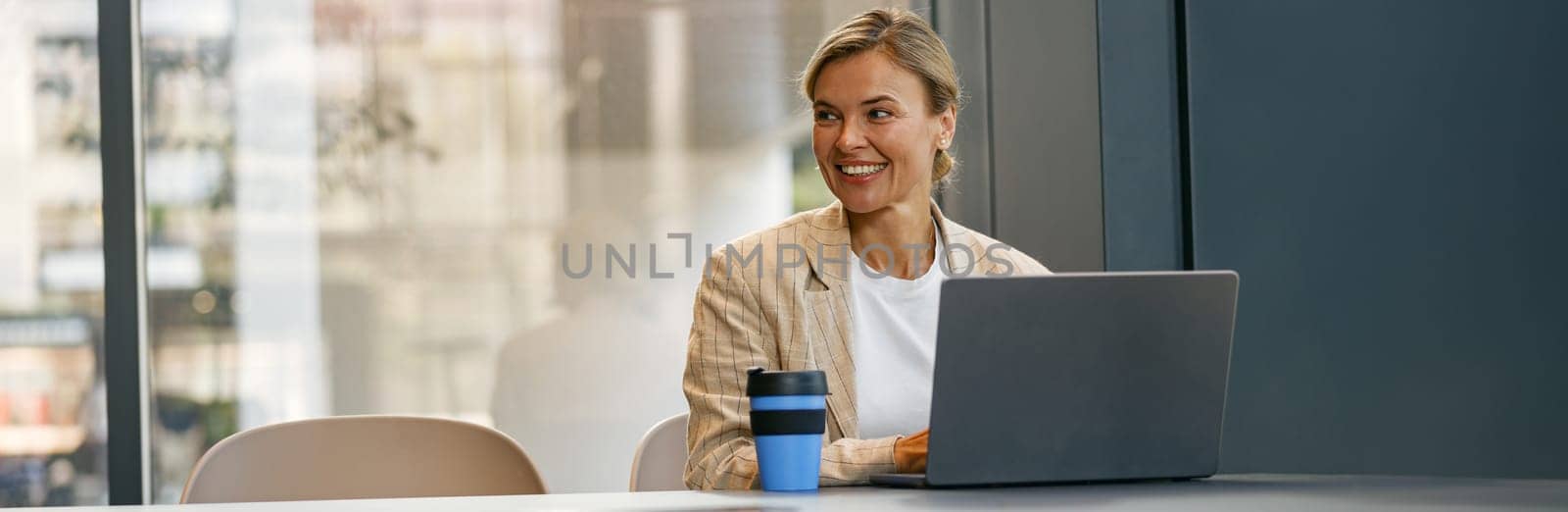 Smiling female entrepreneur working on laptop sitting the desk on office background and looks away by Yaroslav_astakhov
