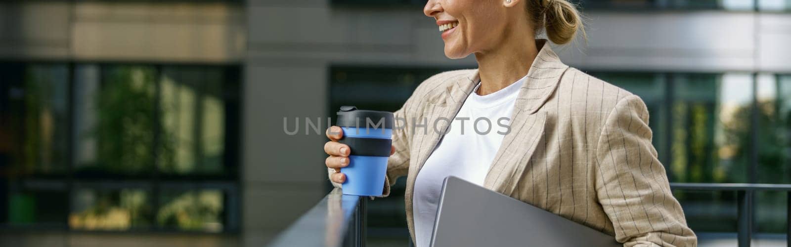 Woman freelancer standing with laptop and coffee cup on modern building background during break time