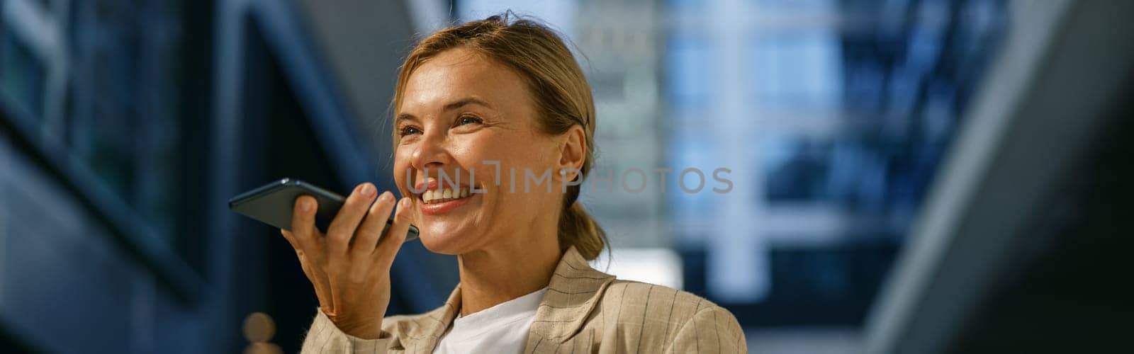 Smiling woman sales manager record audio message to client standing on office building background