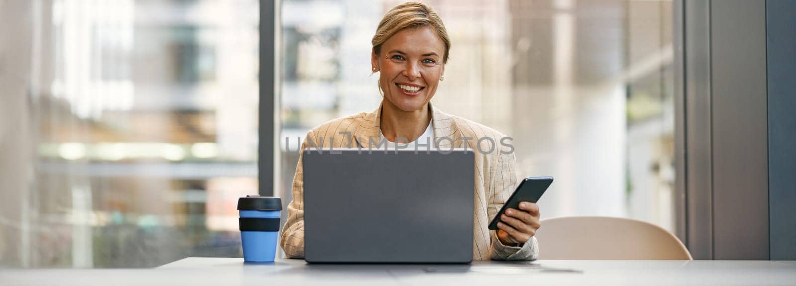 Cheerful female manager use phone while sitting in modern coworking and working on laptop by Yaroslav_astakhov