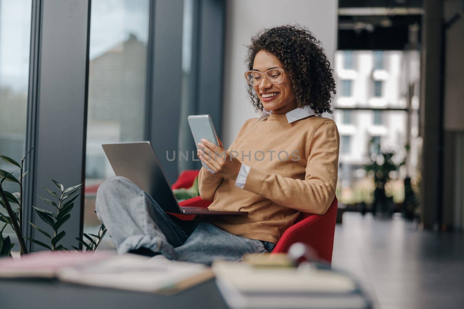 Smiling business woman is using phone sitting in office during break time with laptop