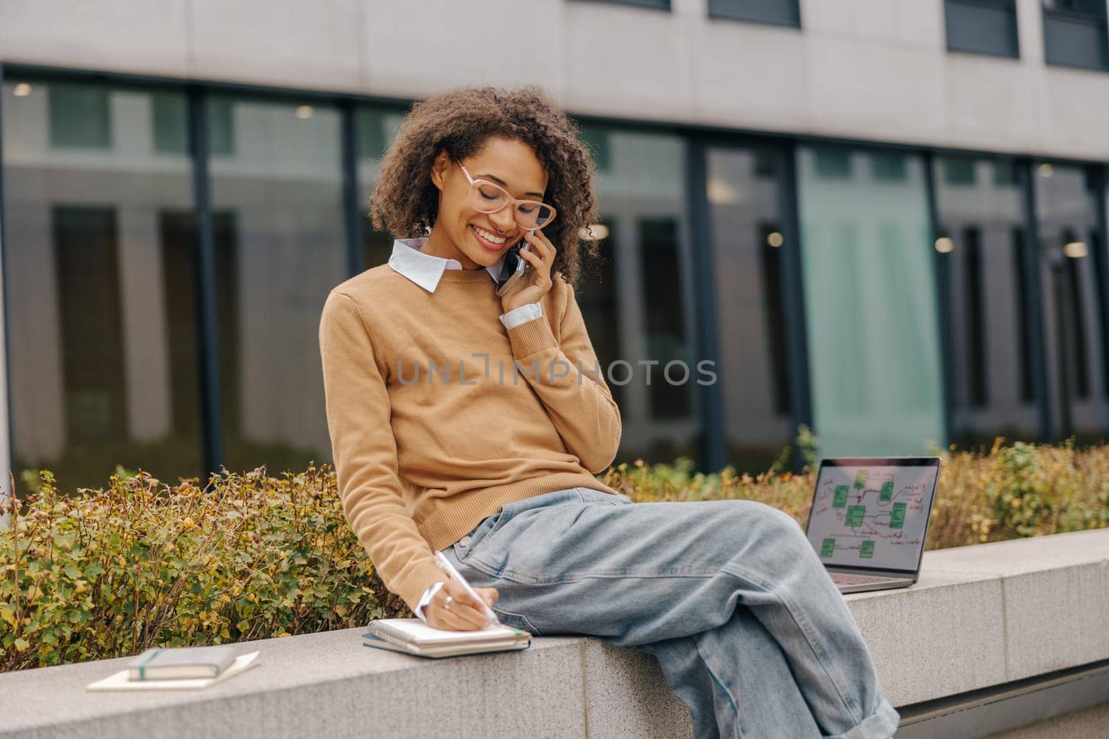 Cheerful business woman talking phone and making notes while sitting on building background