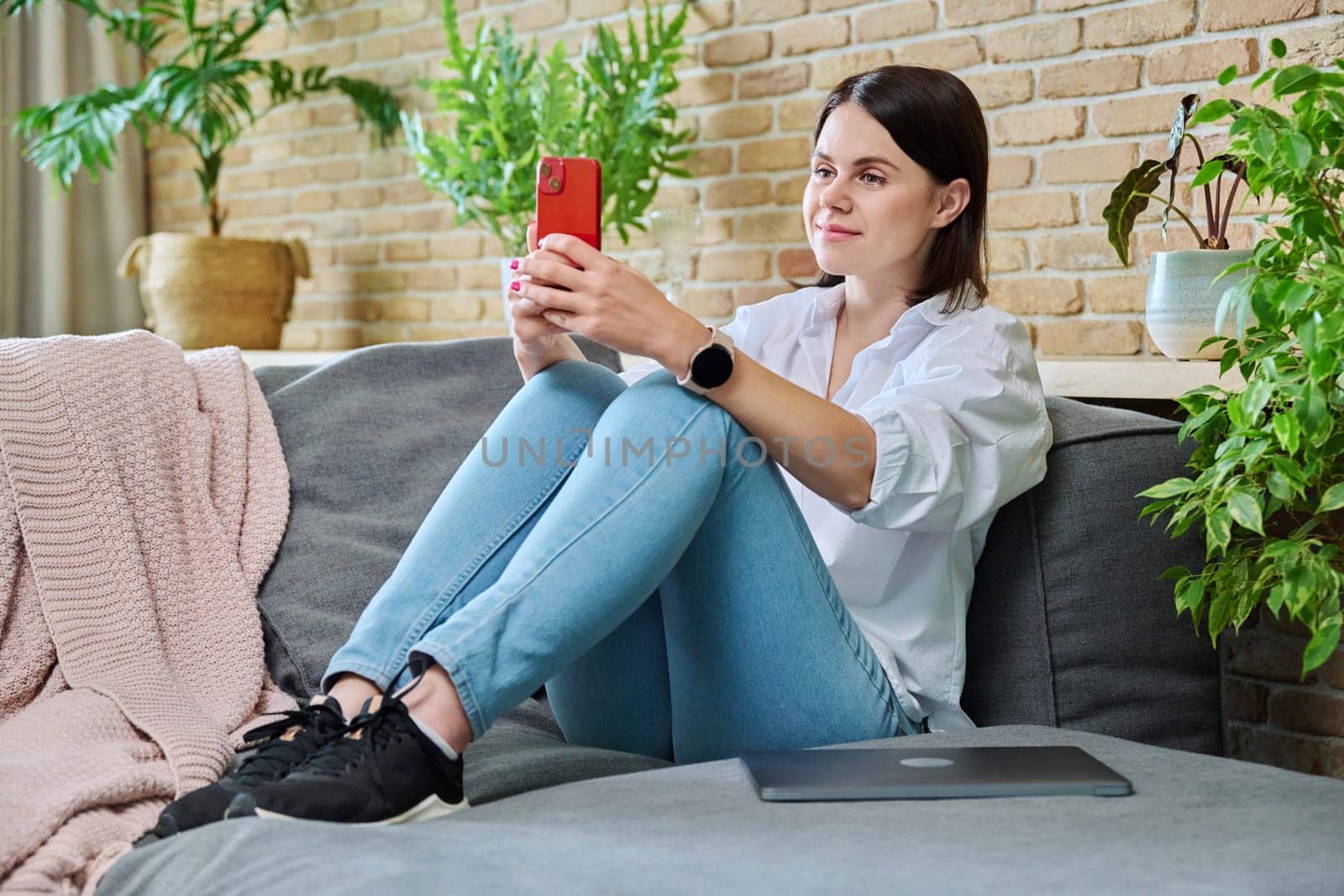 Young smiling happy woman using smartphone, sitting on sofa at home in living room. Internet online services, communication, mobile applications, technologies for leisure study work lifestyle