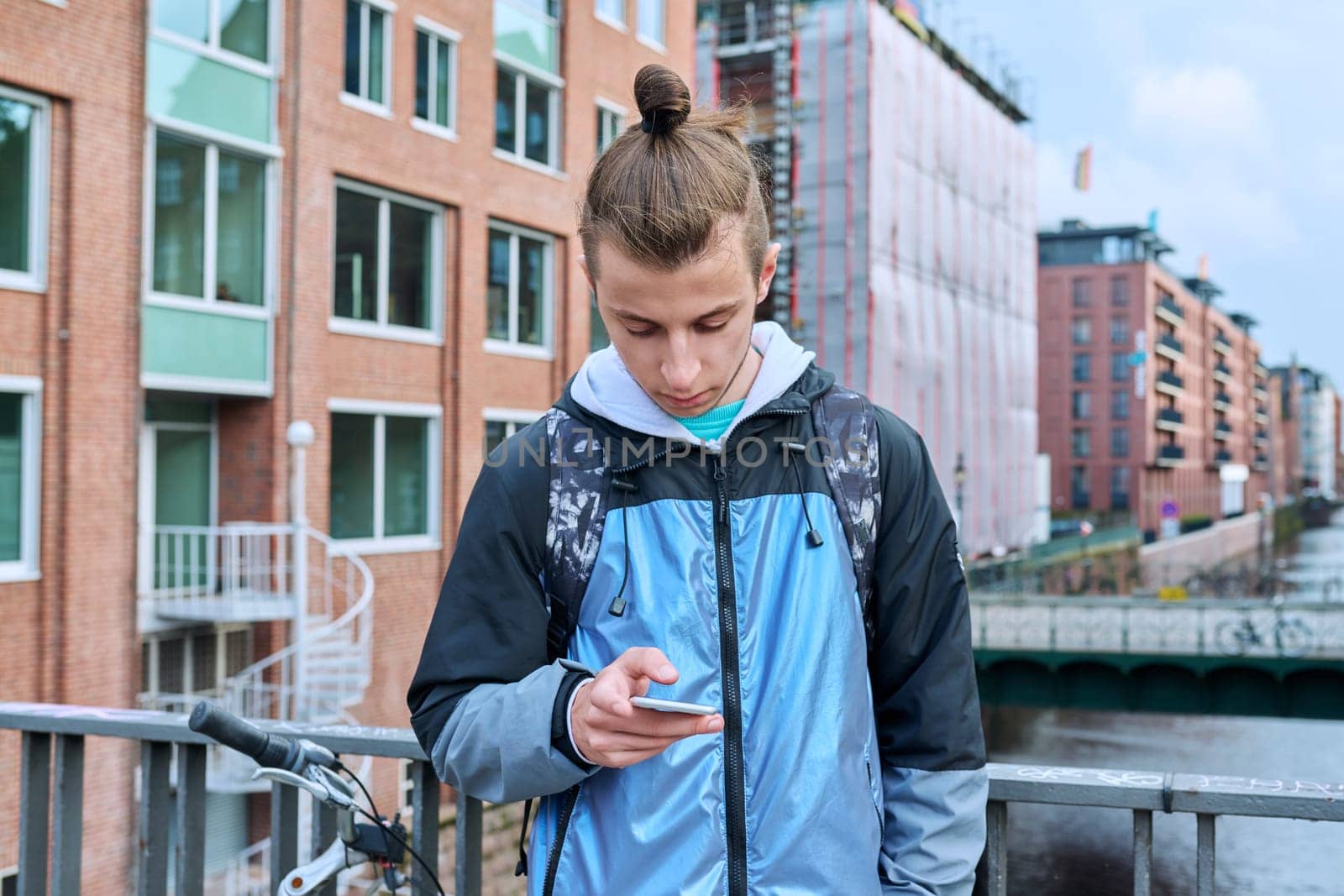 Fashionable young male with backpack using smartphone outdoors in city. Hipster with trendy hairstyle beard with phone in hands mobile applications for tourism travel communication entertainment study