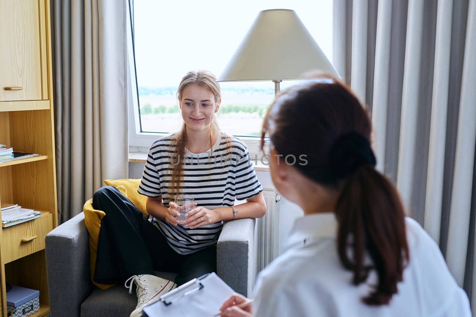 Young female teenager 16, 17 years old talking with counselor psychologist behavior social worker teacher, sitting in chair in office. Psychology psychotherapy therapy mental health adolescence youth