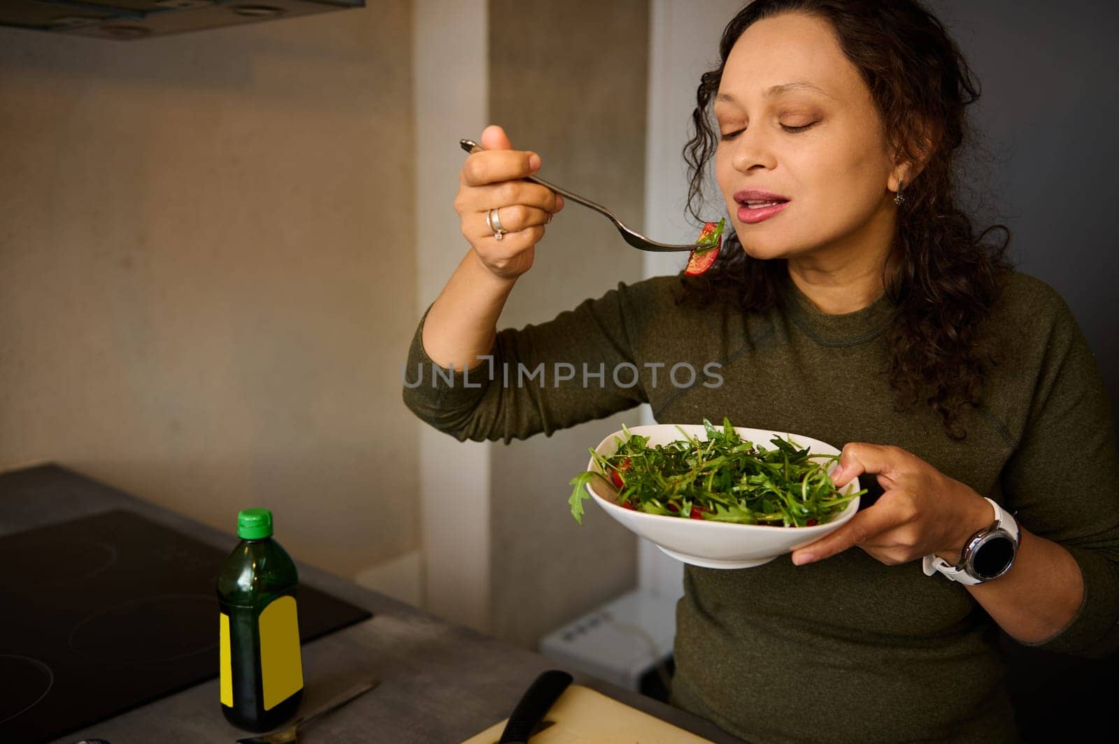 Happy young woman eating healthy salad in the home kitchen. Caucasian pretty female holding a bowl with salad of fresh ripe organic tomatoes and arugula. Healthy eating. Slimming and dieting concept