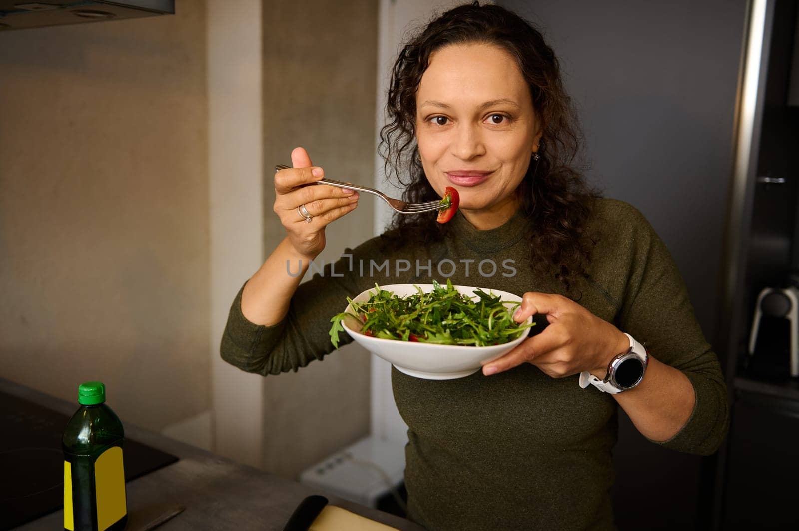 Happy young woman smiles looking at camera while eats healthy salad in the home kitchen by artgf