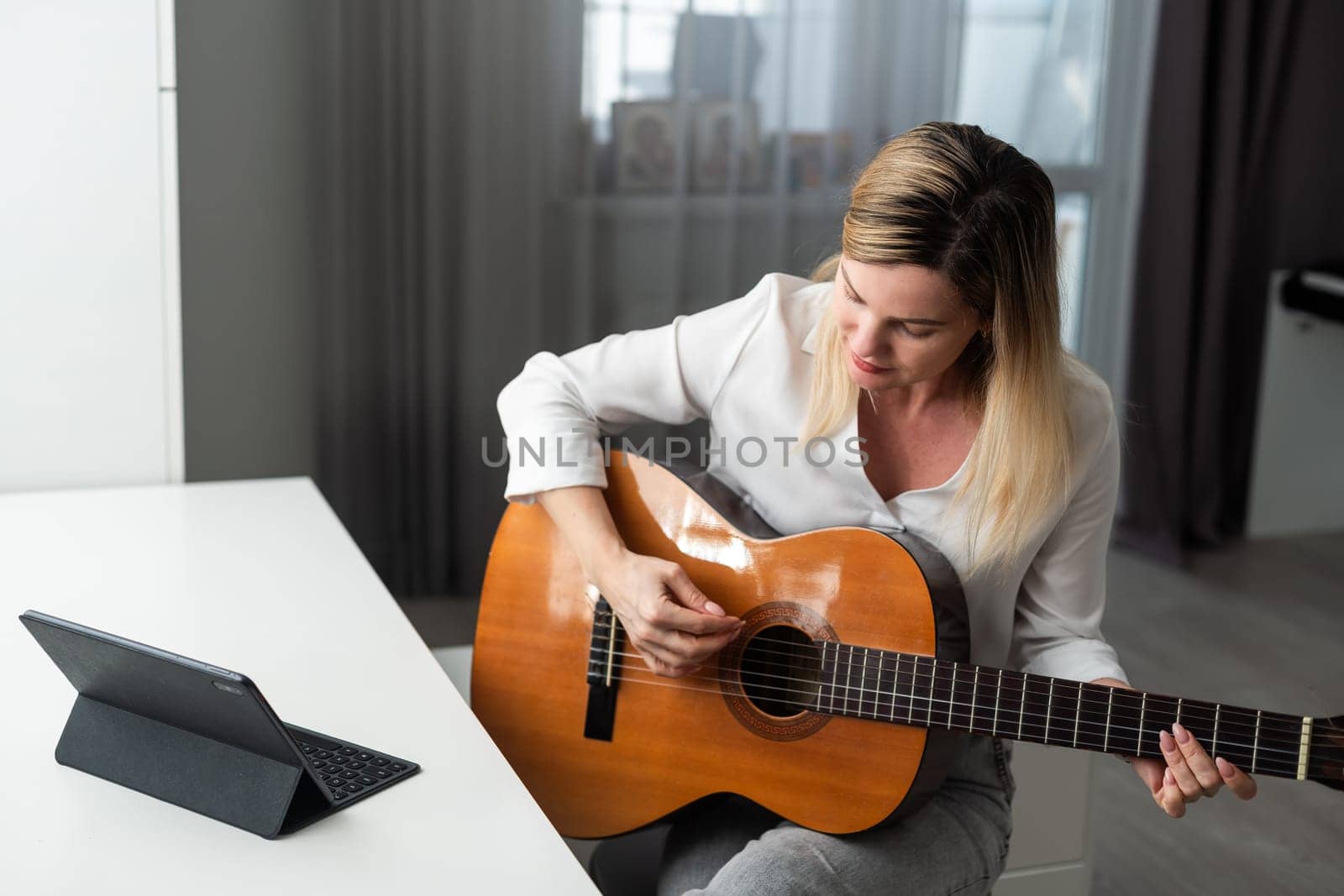 Home Guitar Playing Lesson Girl Hold Instrument. Beautiful Woman at Musical Training. Blonde with Makeup Looking at Notes on Music Stand. Caucasian Girl Guitarist Taking Chords on Strings by Andelov13