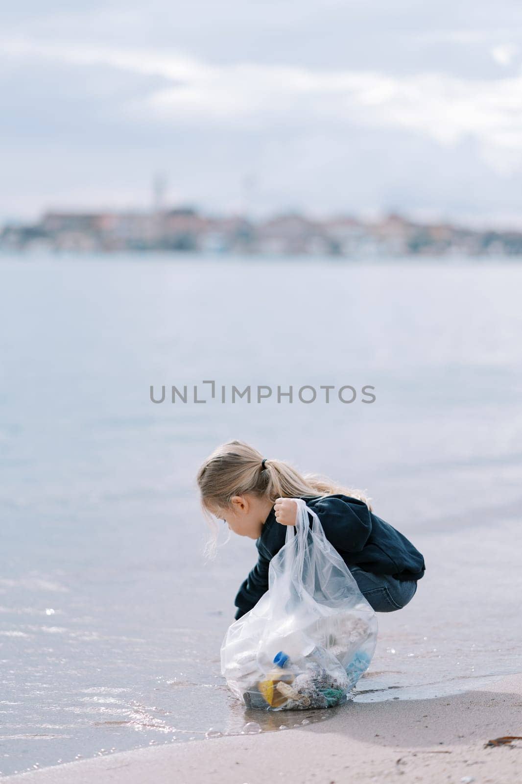 Little girl with a bag squats by the sea and collects garbage. High quality photo