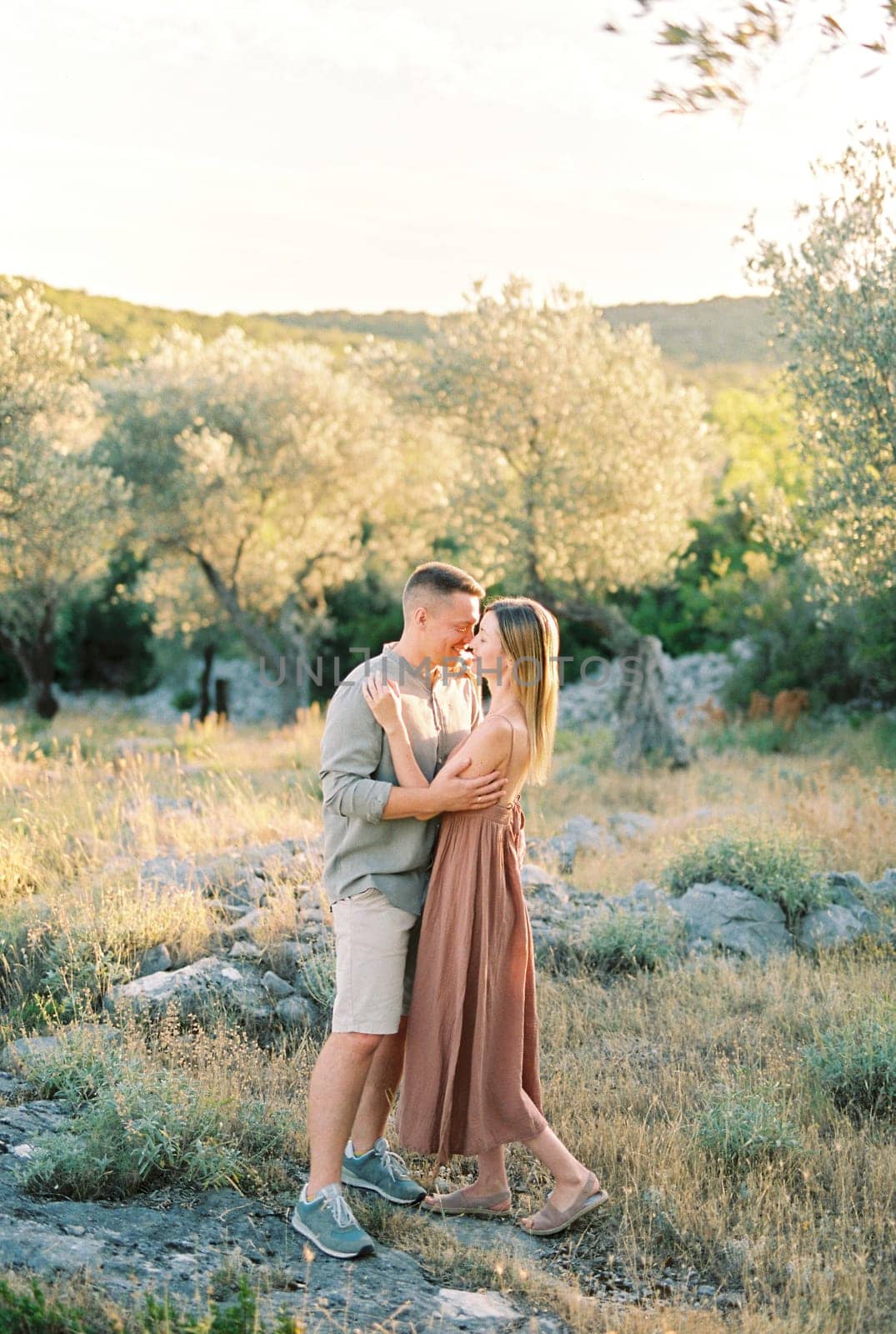 Smiling guy hugs and almost kisses girl while standing in a green garden by Nadtochiy