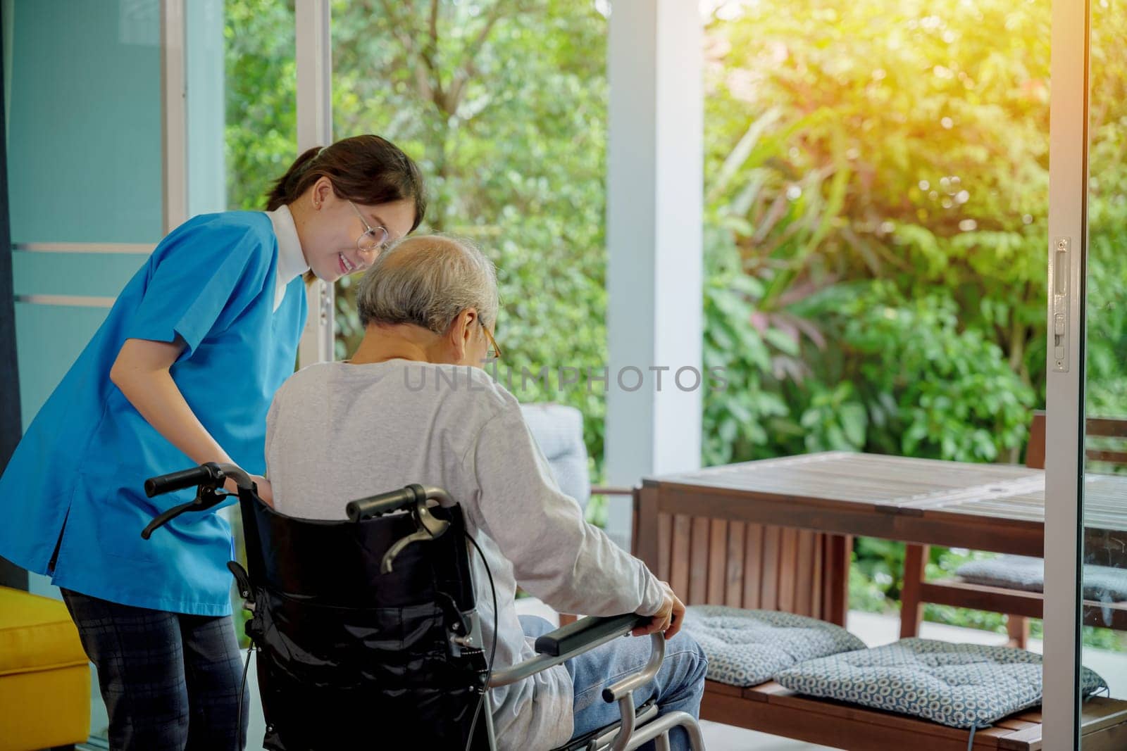 Young Asian nurse is supporting elderly man sitting in wheelchair walking to looking out window on garden and breathe fresh air, pension retirement center, senior man at home, healthcare concept