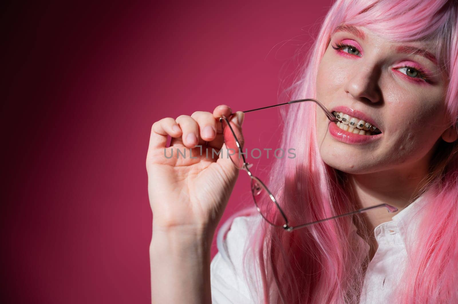 Close-up portrait of a young woman with braces in a pink wig and sunglasses on a pink background