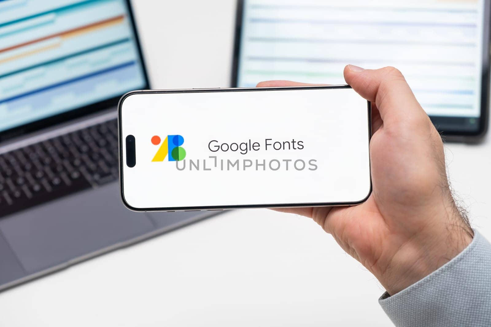Google Fonts application logo on the screen of smart phone in mans hand, laptop and tablet on the table by vladimka