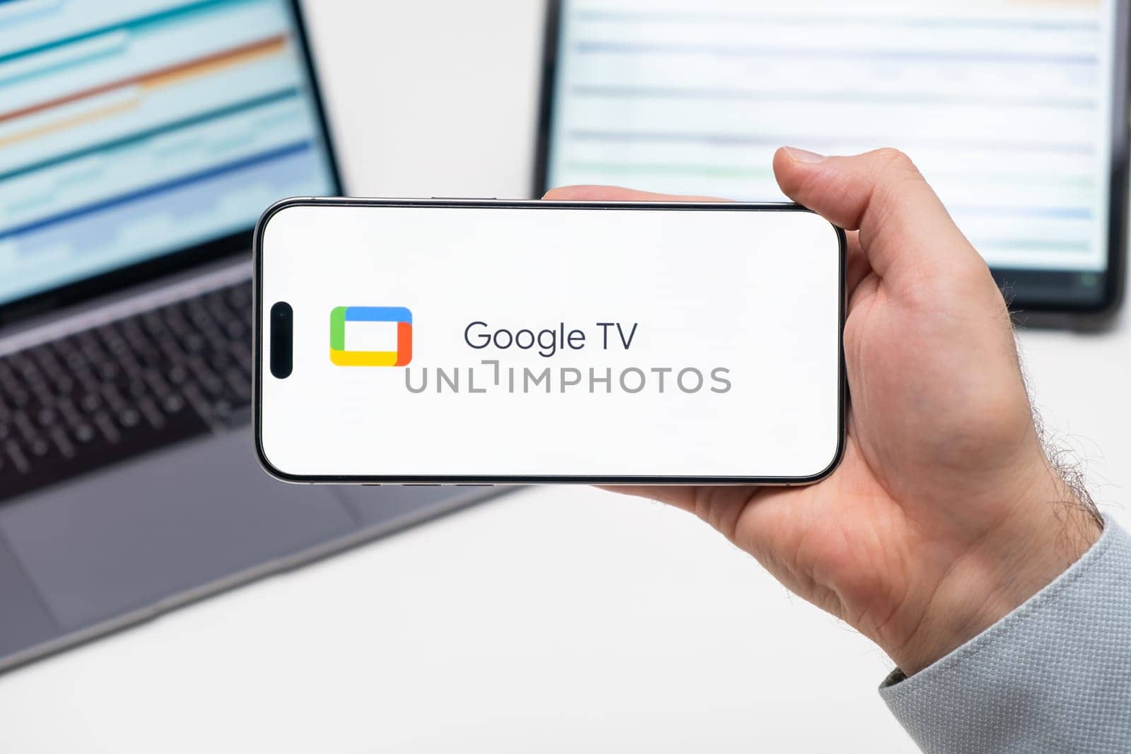 Google TV logo of app on the screen of mobile phone held by man in front of the laptop and tablet by vladimka