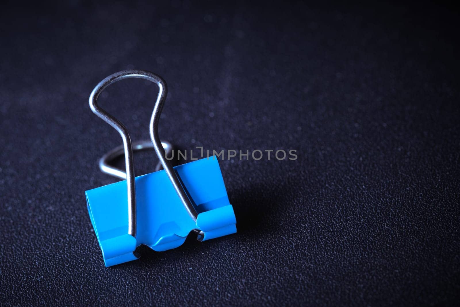 Blue metal paper clip laying on dark blue textured background with copy space. Close-up macro photography, high angle top view