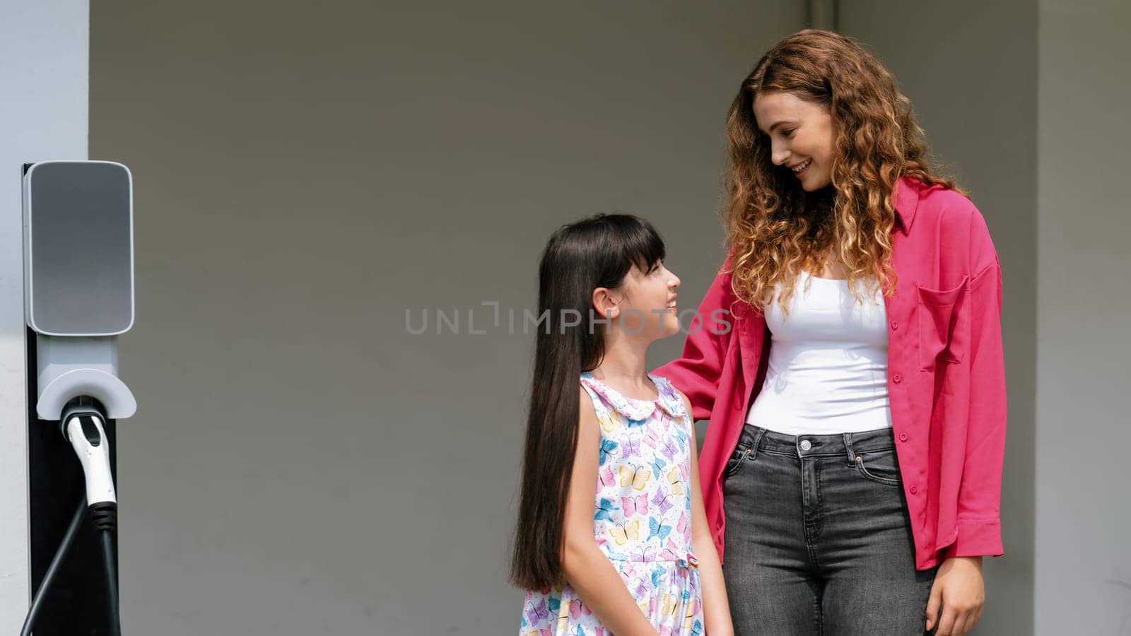 Happy little young girl learn about eco-friendly and energy sustainability as she help her mother recharge electric vehicle from home EV charging station. EV car and modern family. Panorama Synchronos