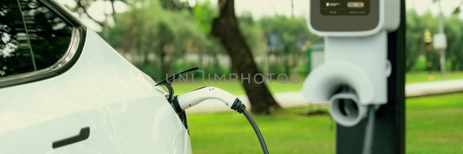 EV electric vehicle recharging battery from EV charging station in outdoor green city park scenic. Natural protection with eco friendly EV car travel. Panorama Banner Exalt