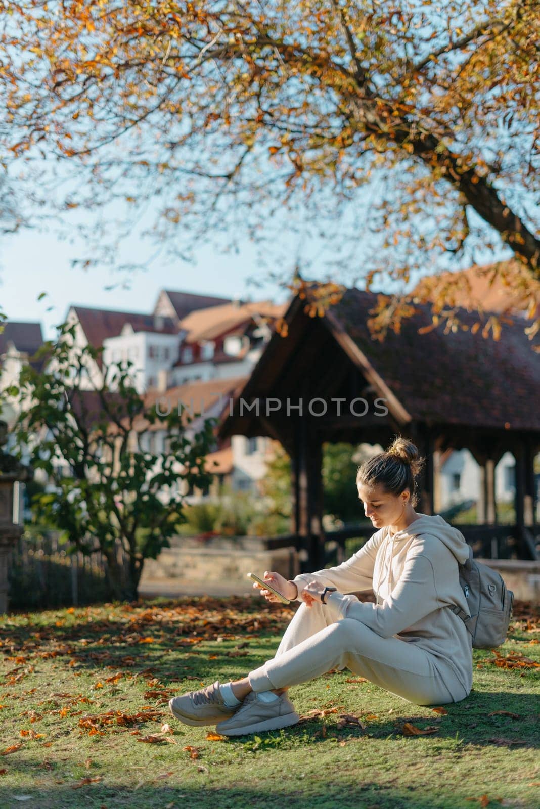 Young Fashionable Teenage Girl With Smartphone In Europian Park In Autumn Sitting At Smiling. Trendy Young Woman In Fall In Park Texting. Retouched, Vibrant Colors. Beautiful Blonde Teenage Girl Wearing Casual Modern Autumn Outfit Sitting In Park In Autumn. Retouched, Vibrant Colors, Brownish Tones. by Andrii_Ko