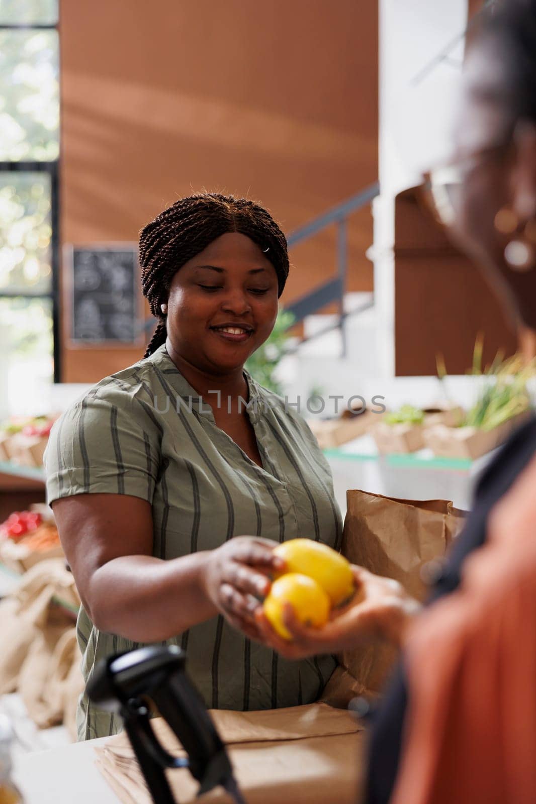 Smiling black woman handing over freshly harvested lemons to vendor for weighing at checkout counter. Portrait of african american consumer with paper bag giving locally grown fresh produce to cashier