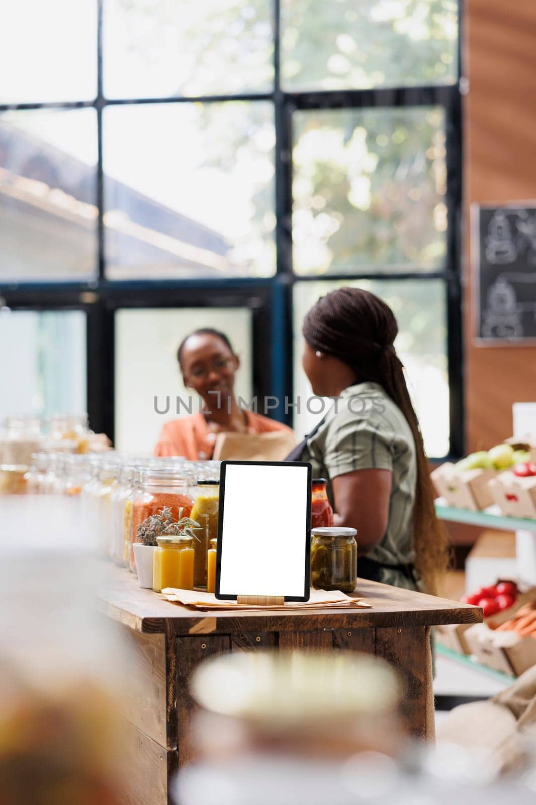 White screen on tablet displays an ad template promoting fresh, organic and sustainable food products in an eco-friendly marketplace. Vendor speaks with client while device shows blank mockup.