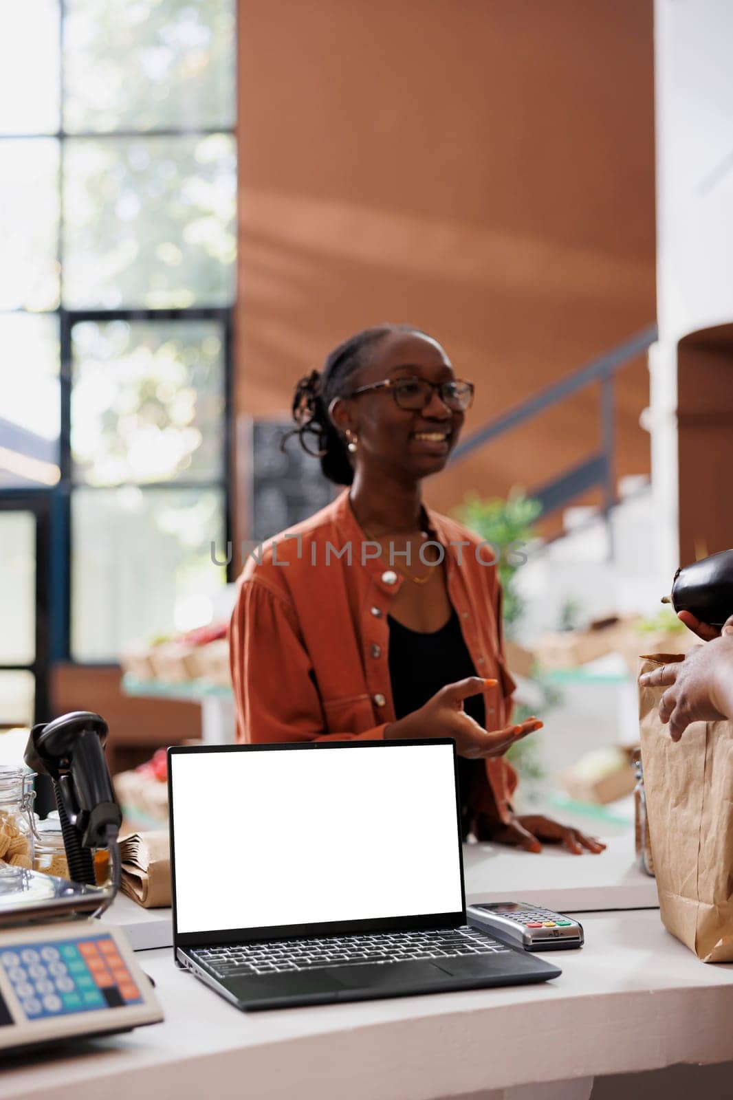 White screen visible on laptop while woman in glasses receives organic products. On counter is a standalone device with copy space, perfect for advertising an environmentally conscious bio food store.