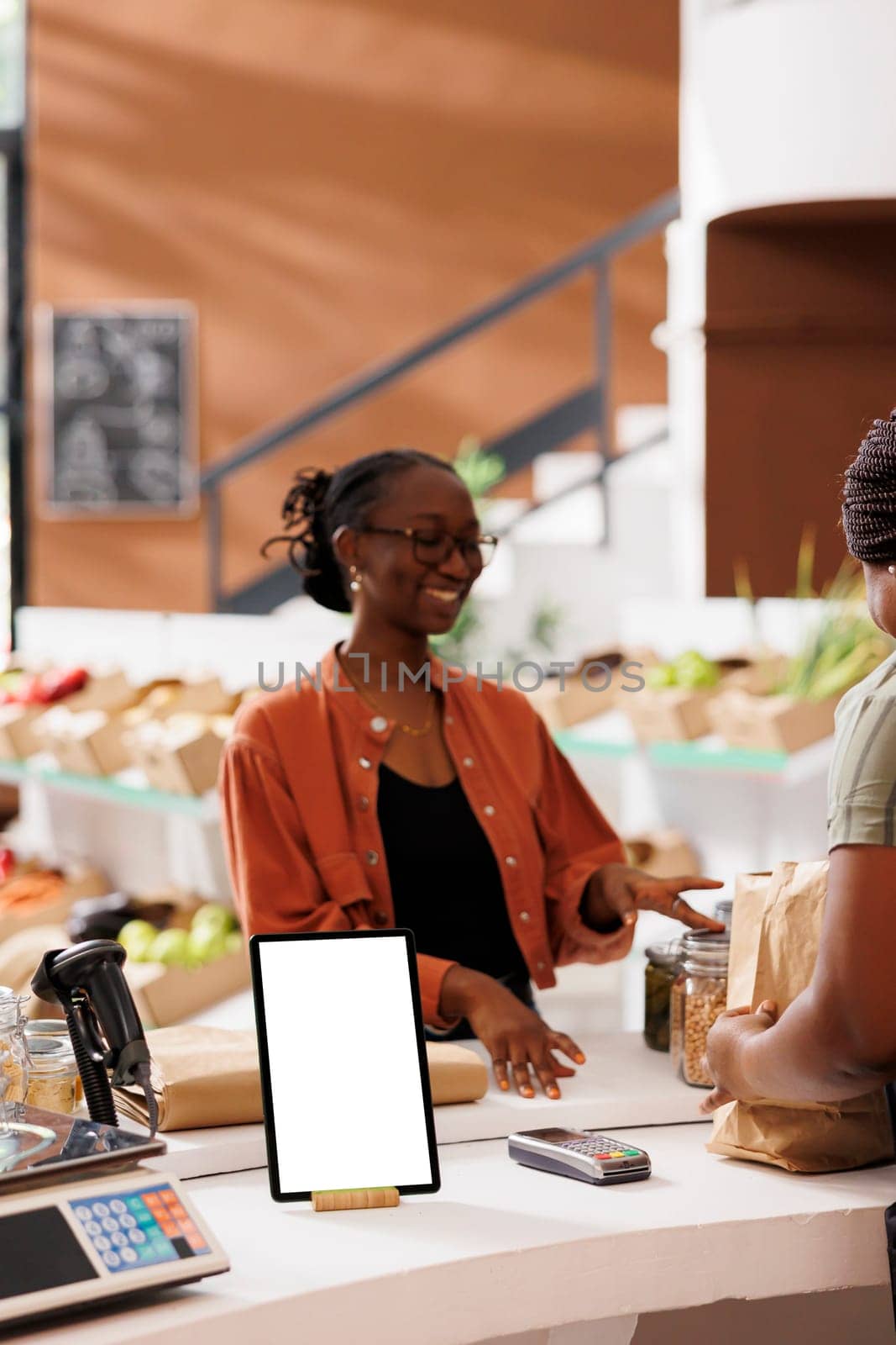 Tablet with blank mockup copy space used for advertising in ethically conscious zero waste supermarket. At store counter, female customer waits for package while digital device displays white screen.