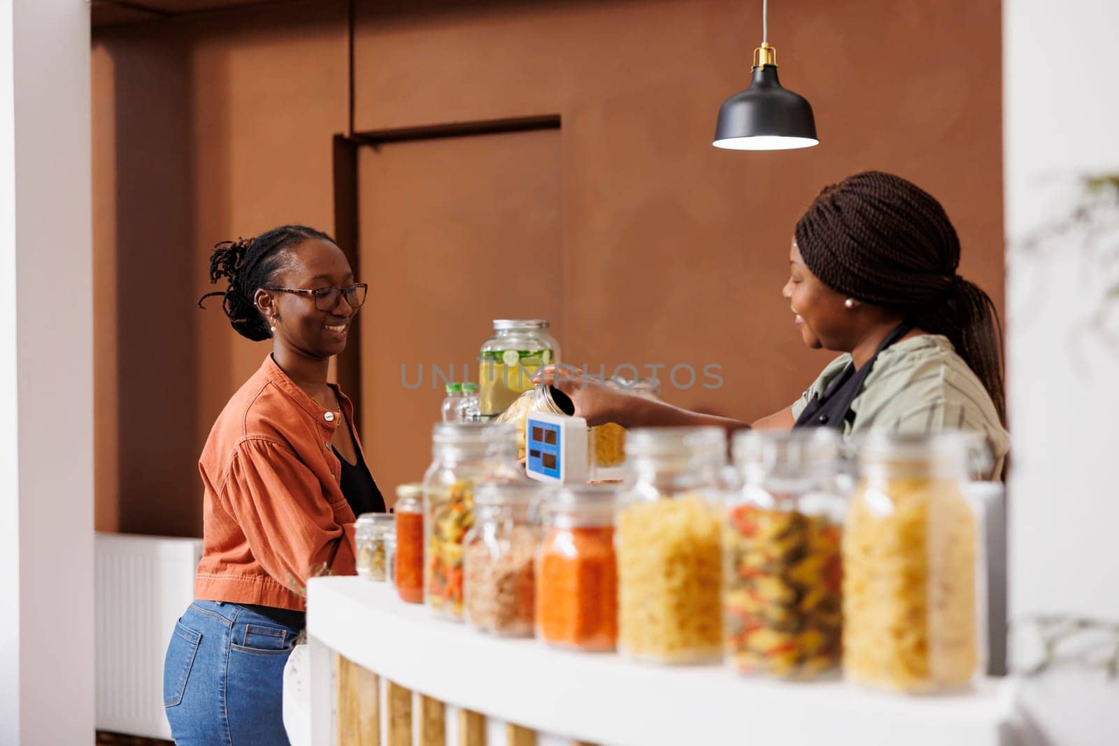 African American woman shopping in eco-friendly supermarket, buying fresh local produce. At checkout counter, black female consumer handing glass jar of pasta to vendor for weighing.