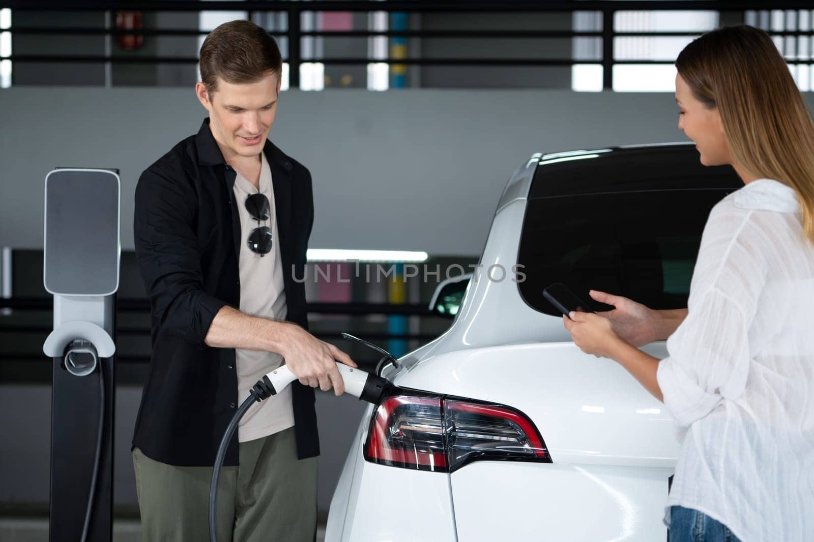 Young couple travel with EV electric car to shopping center parking lot charging in downtown city showing urban sustainability lifestyle by green clean rechargeable energy of electric vehicle innards