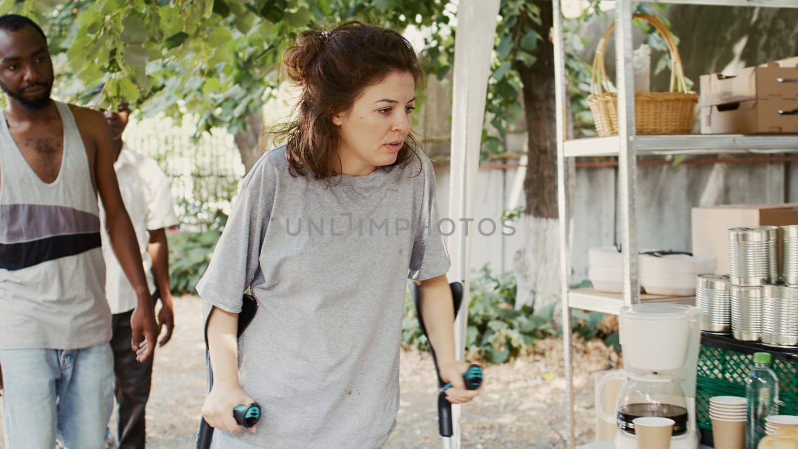 Poor, hungry Caucasian lady on crutches approaches an outdoor food bank to receive free food assisted by black woman. Volunteers serving meals and nourishments to underprivileged and homeless people.