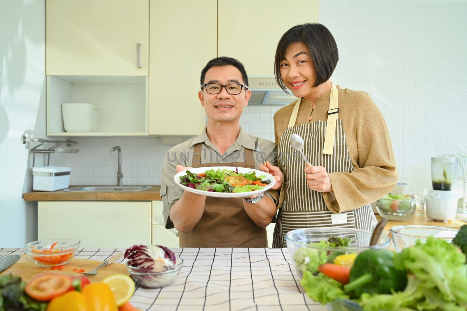 Cheerful retired senior couple preparing healthy vegan salad in kitchen. Healthy eating and culinary concept by prathanchorruangsak